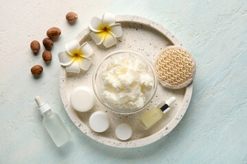Glass bowl of shea butter with bottle of cosmetic oil, plumeria flowers and nuts on light blue background