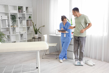 Fototapeta premium Young man measuring his weight on scales and doctor in clinic