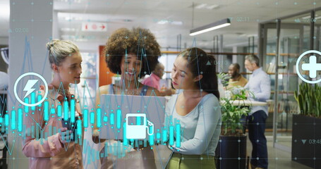 Image of graph and energy icons over diverse female colleagues using laptop at casual office