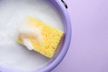 Bucket with foam and sponge on violet background, top view
