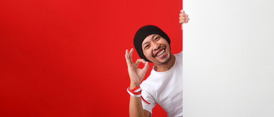 Indonesian man in a white T-shirt and beanie hat shows the OK gesture to the camera behind a white...