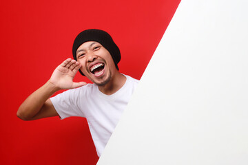 Excited Indonesian man in a white T-shirt and beanie shouts from behind a white wall, announcing a...