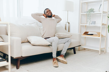 Relaxed African American man sitting on a comfortable sofa at home, smiling and enjoying his weekend He holds a pillow while thinking, with a modern living room as the background This young and