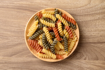 Yellow, orange and green raw fusilli pasta in bowl on wooden background, Food ingredient, Table top view