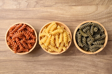 Orange, yellow and green raw fusilli pasta in bowl on wooden background, Food ingredient, Table top view