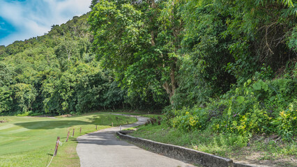 A concrete path winds through a tropical park. Stone and rope fences on the roadsides. Green grass on the lawn. Lush vegetation on the hill. Malaysia. Borneo. Kota Kinabalu.