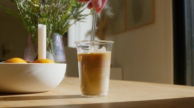 Hand stirring iced latte coffee in a cozy sunlit living room. Refreshing summer drink moment. Perfect for lifestyle or culinary content.