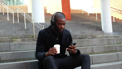black african Businessman in suit and headphones texting on smartphone drinking takeaway coffee - Powered by Adobe