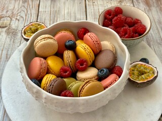 Macarons in different colors and tastes in the white plate and raspberry, blueberry and passion fruit on the marble plate on the wooden background 