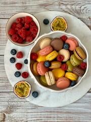 Macarons in different colors and tastes in the white plate and raspberries, blueberries and passion fruit on the marble plate on the wooden background 
