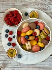 Macarons in different colors and tastes in the white plate and raspberry and blueberry and passion fruit halves on the marble plate on the wooden background 