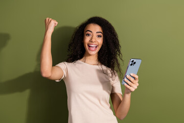 Fototapeta premium Photo of ecstatic girl with wavy hairdo dressed beige t-shirt hold smartphone raising fist win gambling isolated on khaki color background
