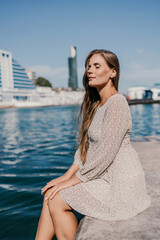 A woman with long hair is sitting on a dock by the water. She is wearing a white dress and she is looking at the water. The scene has a calm and peaceful mood.