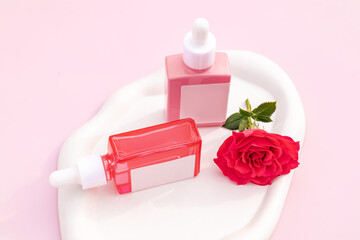 Pink dropper bottles with serum on white tray on pink background with tender red roses. Mock up of natural beauty product based on rose flowers.