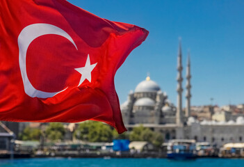 The Turkish flag waves in the wind against blue sky with a mosque and harbour in the background.