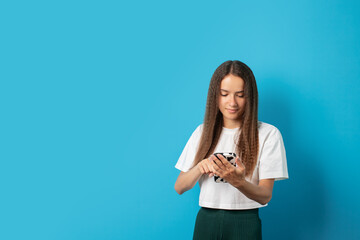 Teenager with mobile phone on blue background, young cute emotional student, using smartphone, smiling at screen, sending a message