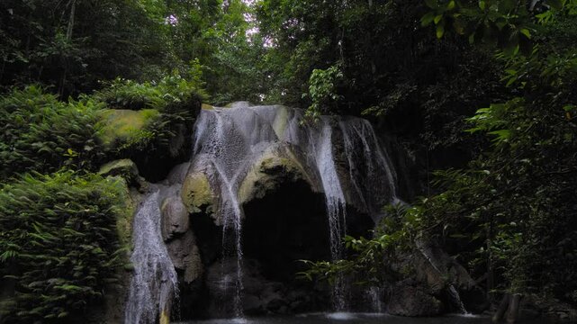 A stunning War Inkabom Waterfall on Batanta Island in Raja Ampat, Indonesia, cascades down a rocky cliff into a serene pool below, surrounded by lush greenery. Tilt-down shot in slow motion.