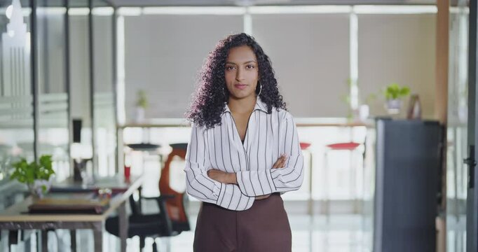 Indian Asian curly hair young adult gen z cute teen age expert girl cross arms standing proud look cam indoor office day happy woman lady staff agent fold hand do pose head shot video job work place	