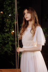 A caucasian woman in an elegant white lace dress sits on a wooden swing amidst dark foliage.