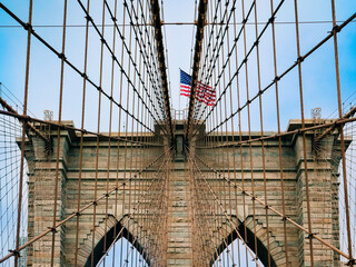 Fototapeta premium Brooklyn Bridge in New York with the US flag flying on a stone pylon. Hanging cables are visible, creating a geometric pattern, and the stamped year 1875. Clear blue in the sky background.