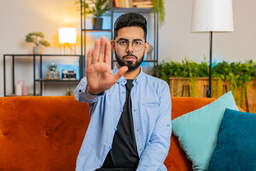 Portrait of Indian Arabian young man guy on sofa with serious expression doing stop gesture with...