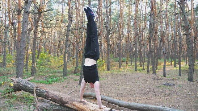 Young man doing handstand on log in forest. Hardy and strong guy doing stunts at wood. Athlete showing performance outdoor. Sportive and healthy lifestyle. Scenic background. Dolly shot