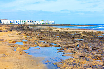 Scenic sandy beach with turquoise ocean waves. Beach with a rocky shoreline and a Famara small town in Lanzarote, Canary Islands
