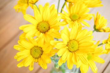 Close up photo of a bunch of yellow daisy flower bouquet in transparent glass vase on wooden table. Modern interior design of modern office work room or workplace with plant.