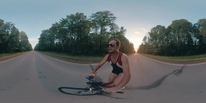 VR 360 footage of the man doing cycling work out. Young man cycling on an asphalt road during sunset