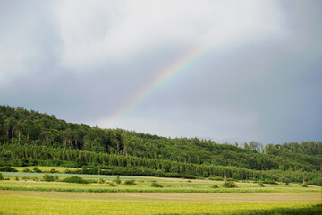 Tęcza nad górami, zielone pola i las na wzgórzu.