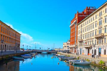 Fototapeta premium Boats cruising along a scenic canal, surrounded by lush greenery and historic buildings in Trieste Italy. Water canal with a row of boats and a bridge 