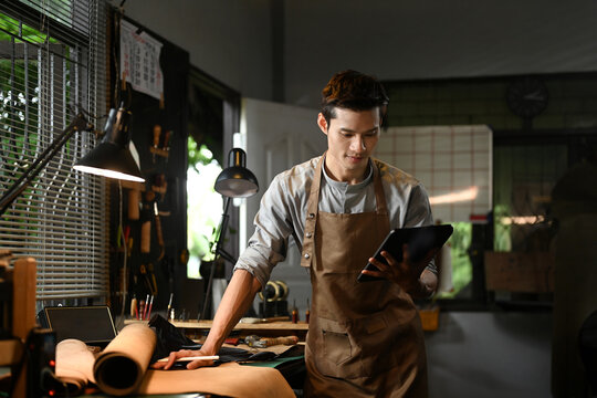Smiling asian male artisan wearing an apron using a digital tablet in his workshop. Small business and technology concept