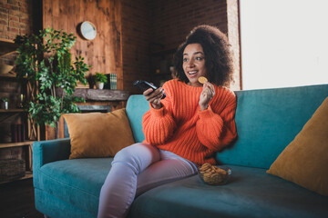 Photo of pretty cheerful lady wear orange sweater watching tv eating snacks having rest indoors room home house