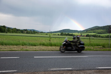 Motocykl na drodze z pasażerką, zielone pola, dachy domu, lasy na wzgórzu. © Stanisław Błachowicz