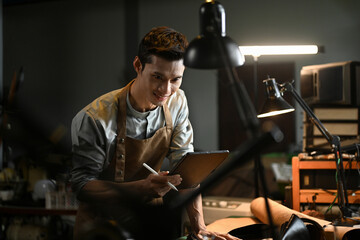 Smiling asian male artisan wearing an apron using a digital tablet in his workshop. Small business and technology concept