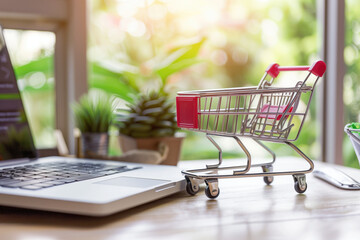 Miniature shopping cart on wooden desk with laptop and green plants in the background. E-commerce, online shopping, digital marketing, retail business, online sales concept.