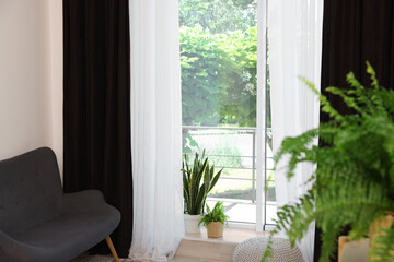 Shelving unit, sofa, green houseplants, pouffe and elegant curtains in room. Interior design