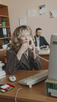 Vertical side shot of stylish female Caucasian secretary in glasses and jacket working on large monitor and picking up stationary phone, discussing work issues at desk in retro office, 90s