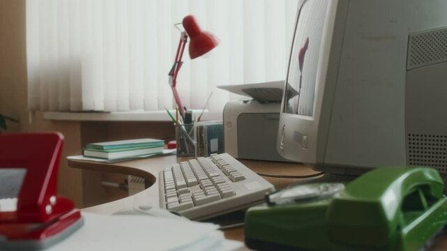 No people side shot of empty workplace for 90s office worker with large computer monitor on desk next to keyboard, printer, green rotary phone, red table lamp and hole puncher on documents, vintage