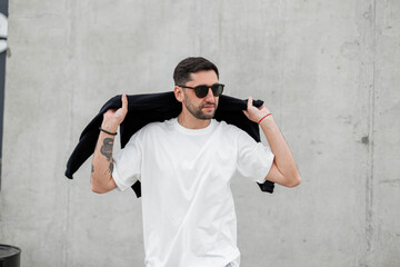 handsome fashion hipster man with sunglasses in a fashionable white T-shirt puts a sweater on his shoulders and walks in the city near a concrete building