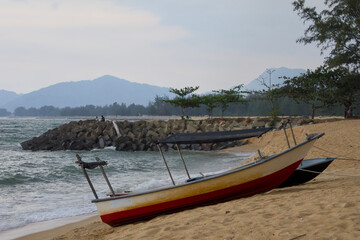 Fototapeta premium A vibrant traditional boat rests on the sandy shore of a tropical beach, framed by hazy mountains in the background at Pantai Teluk Lipat, Dungun, Terengganu.