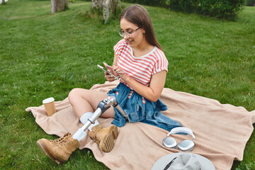 A young woman with a prosthetic leg relaxes on a blanket in a park, scrolling on her phone.