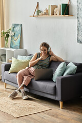Obraz premium A young woman with a prosthetic leg sits on a couch in her living room, wearing headphones and smiling as she uses her laptop.