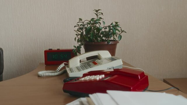 No people tilt shot of two white and red vintage stationary telephones on wooden desk next to digital clock and ream of documents in retro office, 90s