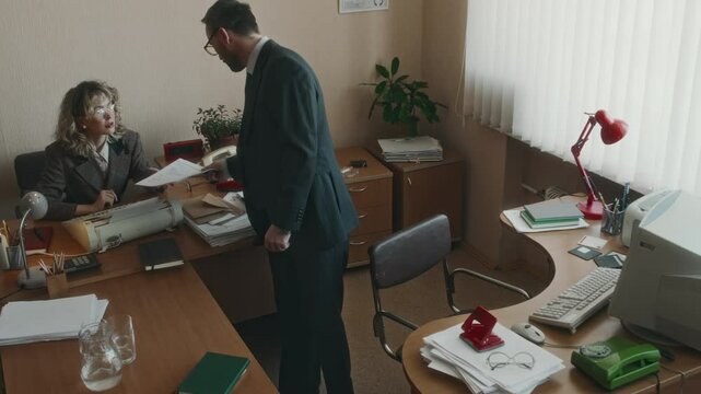 High angle full shot of male Caucasian office worker bringing paperwork to female coworker with typewriter and taking seat, working on old computer in cozy retro office with plants