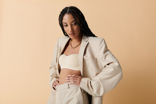 A young woman with long braids poses confidently in a beige blazer and pants, looking down thoughtfully against a neutral backdrop.