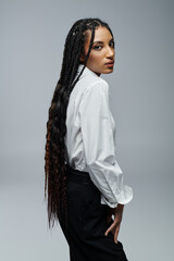Naklejka premium A stylish young woman with long braids poses in a crisp white shirt against a gray backdrop.