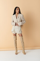 African American woman in long braids poses in stylish beige blazer, skirt and knee-high boots against neutral background