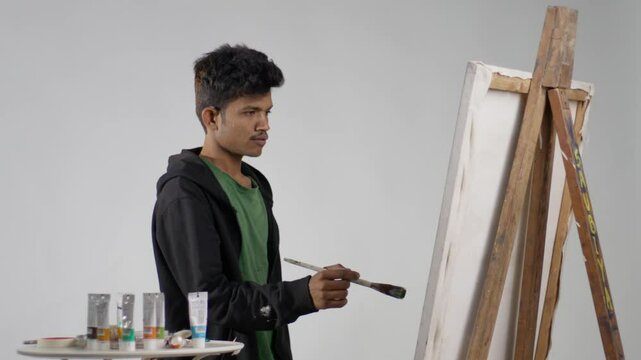 A static mid shot of a man painting on a canvas board kept on an easel and paint colours by this side, with his shadow on the white wall