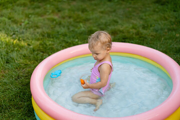 Baby in a pink swimsuit is swimming in an inflatable pool and eating an apricot in the backyard. 